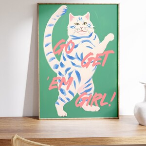 May include: A white cat with blue stripes stands on a green background. The cat is raising its right paw and has a playful expression. The text "GO GET 'EM GIRL!" is written in pink on the green background.