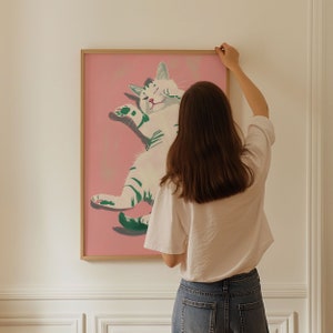 May include: A framed illustration of a white cat with green stripes on a pink background. The cat is lying on its back with its paws in the air. A person is hanging the framed illustration on a white wall.