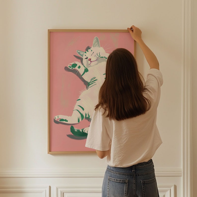May include: A framed illustration of a white cat with green stripes on a pink background. The cat is lying on its back with its paws in the air. A person is hanging the framed illustration on a white wall.