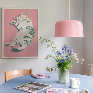 May include: A light blue table with a white cat illustration in a pink frame, a vase of blue and white flowers, a pink pendant light, and a white mug on the table.