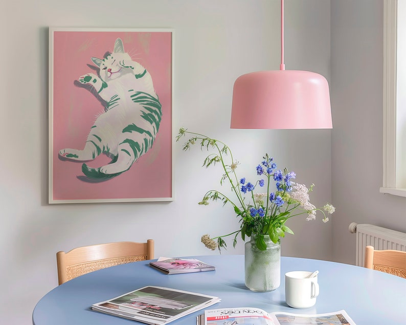 May include: A light blue table with a white cat illustration in a pink frame, a vase of blue and white flowers, a pink pendant light, and a white mug on the table.