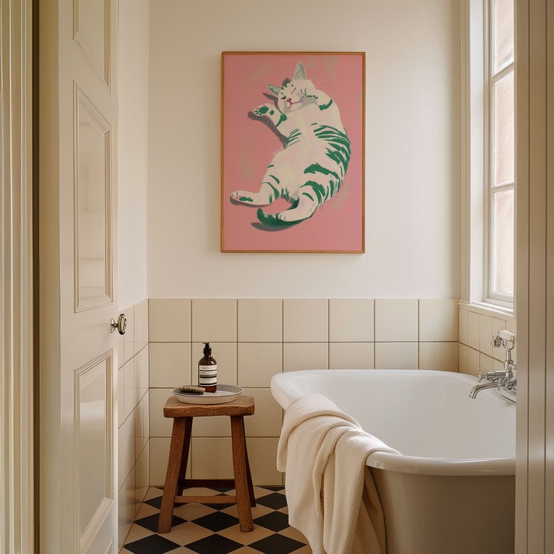 May include: A white and green cat with closed eyes is depicted in a framed painting hanging on a white tiled bathroom wall. The cat is lying on its back with its paws in the air. The painting is on a pink background.