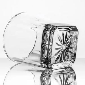 May include: A clear glass tumbler with a square base and a starburst pattern on the bottom. The glass is empty and has a slight reflection.