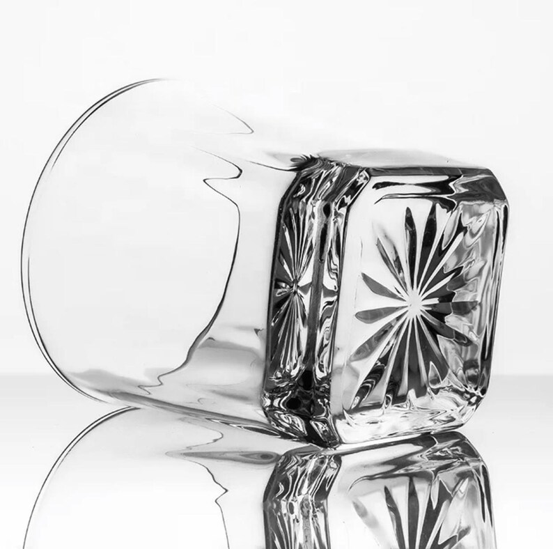 May include: A clear glass tumbler with a square base and a starburst pattern on the bottom. The glass is empty and has a slight reflection.
