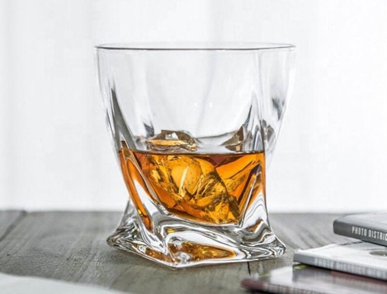 May include: A clear glass whiskey tumbler with a unique, twisted design. The glass is filled with amber-colored whiskey and ice cubes.