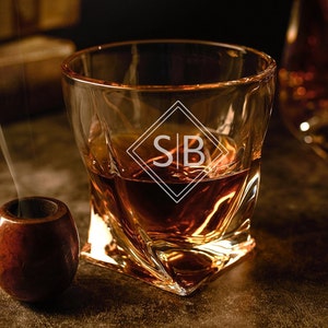May include: A clear glass rocks glass with a twisted design, filled with amber liquid. The glass has a diamond-shaped logo with the initials "S|B" on it. A wooden pipe sits on a dark surface next to the glass.