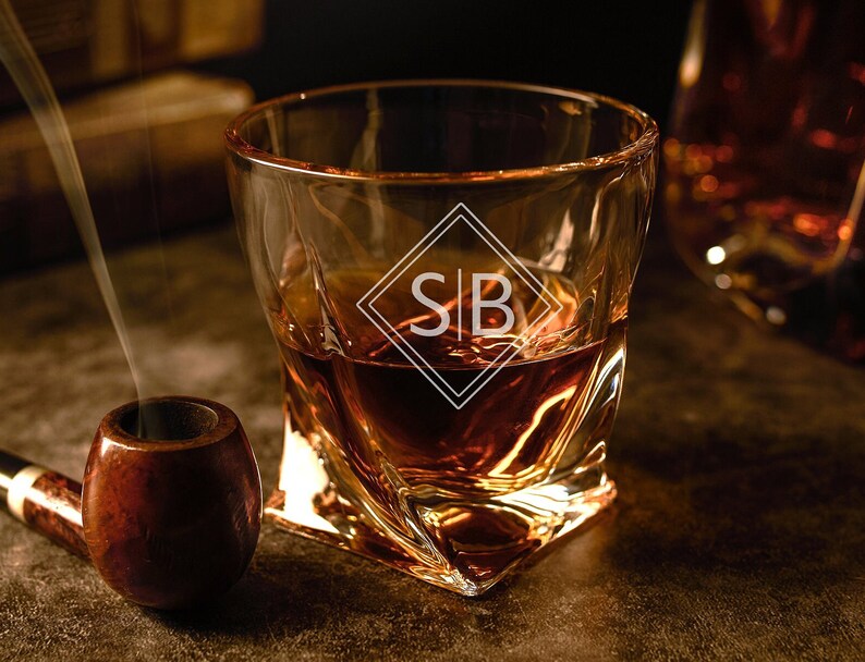 May include: A clear glass rocks glass with a twisted design, filled with amber liquid. The glass has a diamond-shaped logo with the initials "S|B" on it. A wooden pipe sits on a dark surface next to the glass.