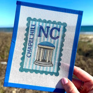 Puede incluir: Un bordado azul y blanco con un diseño de Chapel Hill, NC. La obra de arte tiene forma de sello con las letras "NC" y "Chapel Hill" visibles. La pieza se sostiene contra un fondo de cielo azul y playa.