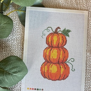 May include: A needlepoint canvas featuring a stack of three orange pumpkins with a brown stem and green leaves. The design is printed on a white grid mesh fabric with a small colour palette guide at the bottom showing yellow, orange, red, brown, and green.