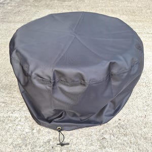 May include: A dark grey, round outdoor furniture cover. The cover is made of a smooth, water-resistant material and features a drawstring closure at the bottom. Designed to protect outdoor furniture from the elements.