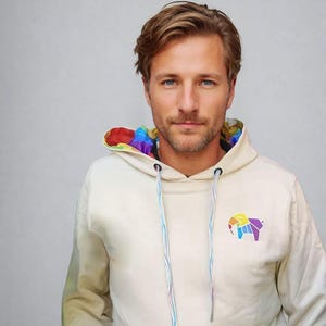 Puede incluir: Una sudadera con capucha de color crema con un forro interior arcoíris y un diseño de elefante geométrico de colores en la parte delantera.