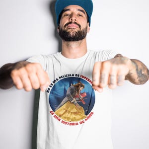Op de afbeelding: Wit T-shirt met een ronde afbeelding van een man en een vrouw in een kasteelomgeving. De tekst rond de afbeelding luidt "No es una pelicula de superheroes, es una historia de amor."