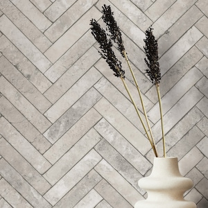 Puede incluir: Un jarrón de cerámica blanco con un diseño ondulado texturizado contiene tres tallos de flores secas con cabezas de semillas de color marrón oscuro. El jarrón está delante de una pared con un patrón de espiga de azulejos de color gris claro.