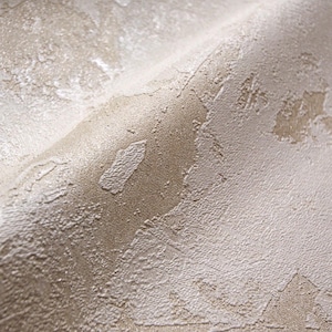 Puede incluir: Primer plano de papel pintado texturizado con un aspecto envejecido y moteado. La superficie presenta una mezcla de tonos beige y blanquecino, con patrones irregulares en relieve que crean una estética táctil y envejecida. El papel pintado tiene un brillo sutil.