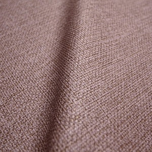 May include: Close-up of a textured fabric in a muted mauve color. The weave is visible, creating a subtle pattern. The fabric has a soft sheen, suggesting a blend of materials. The overall appearance is smooth and inviting.