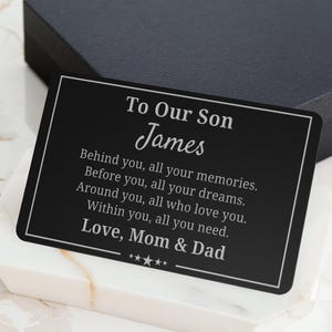 May include: A black rectangular card with white text, resting on a white and gray marble-patterned surface. The card reads "To Our Son James" followed by a heartfelt message from "Mom & Dad."