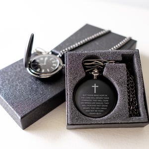 May include: A black pocket watch with a silver chain and a black box. The watch face has a white cross and the text "But those who hope in the Lord will renew their strength, they will soar on wings like eagles: they will run and not grow weary, they will walk and not be faint. - Isaiah 40:31"