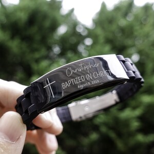 K&ouml;nnte beinhalten: Schwarzes Silikonarmband mit einer silbernen Metallplatte, auf der "Christopher Baptized in Christ August 8, 2024" und ein Kreuz eingraviert sind.