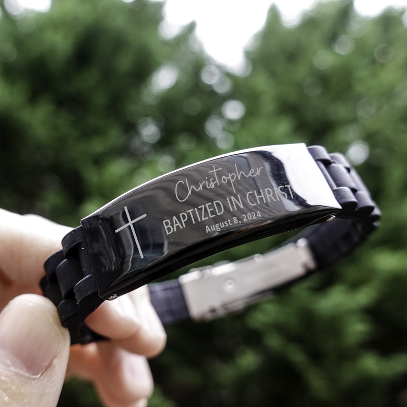 K&ouml;nnte beinhalten: Schwarzes Silikonarmband mit einer silbernen Metallplatte, auf der "Christopher Baptized in Christ August 8, 2024" und ein Kreuz eingraviert sind.
