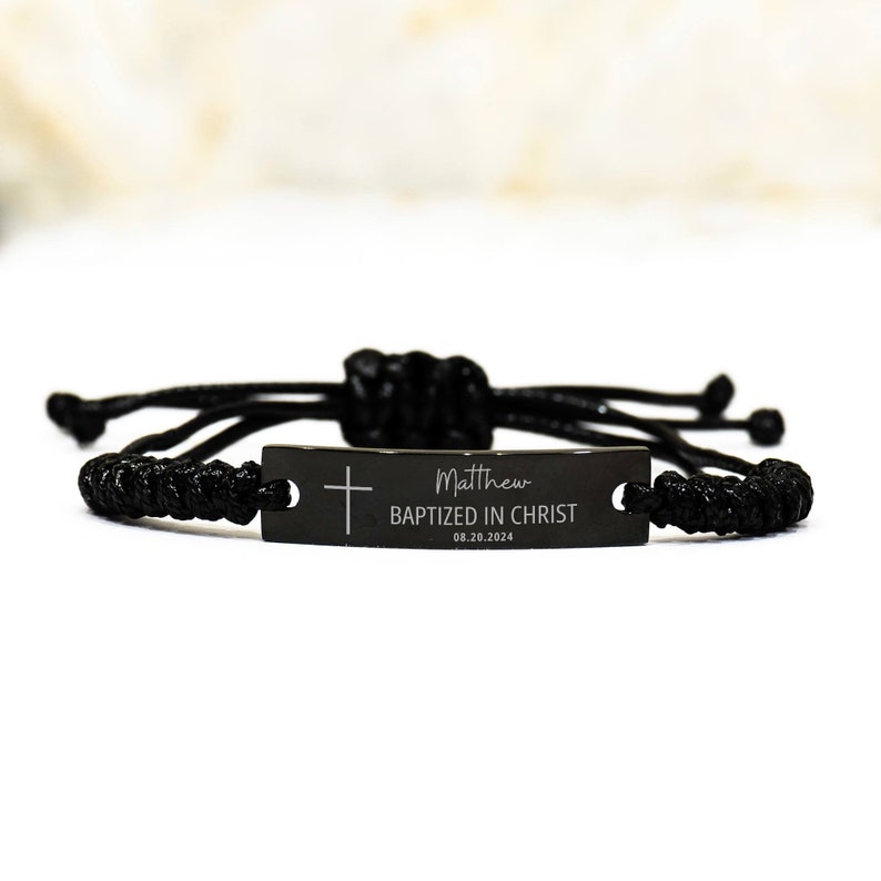K&ouml;nnte beinhalten: Schwarzes geflochtenes Armband mit einem schwarzen Metalletikett, auf dem "Matthew Baptized in Christ 08.20.2024" und ein kleines Kreuz stehen.