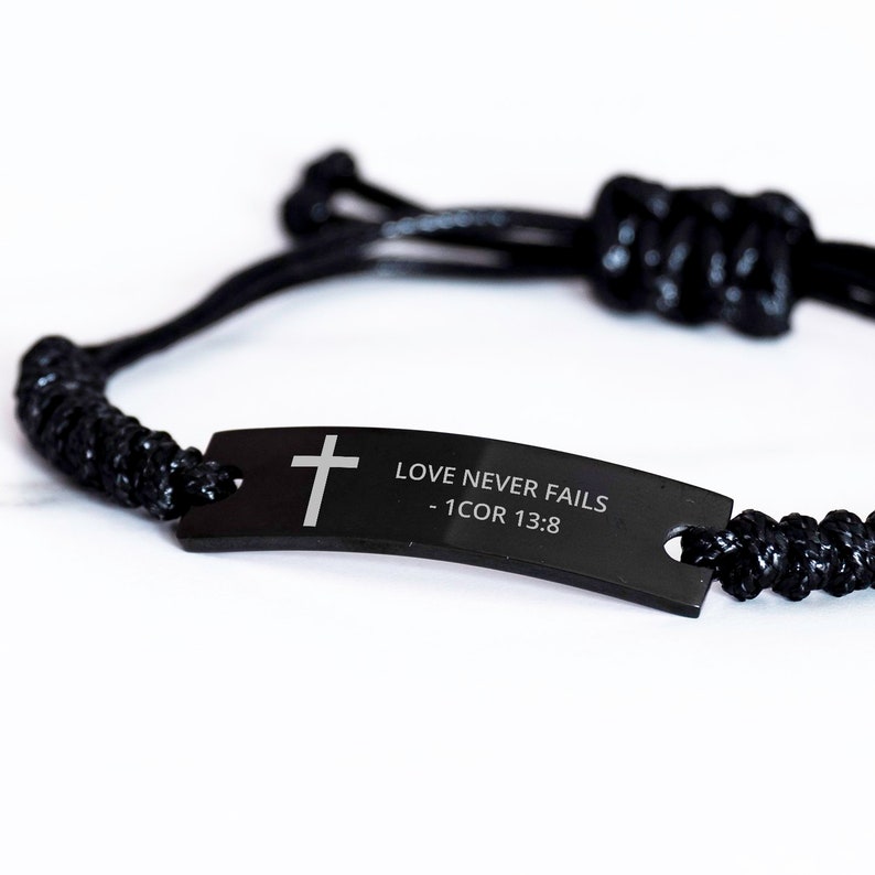 May include: Black braided bracelet with a black metal tag that reads "LOVE NEVER FAILS - 1 COR 13:8" and features a silver cross.