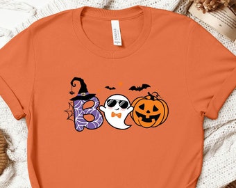 Può includere: Maglietta arancione con una grafica di un fantasma con occhiali da sole e un papillon, un cappello da strega e una zucca. Il testo "BOO" è scritto con gli elementi grafici.