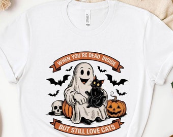 以下が含まれることがあります： 黒い猫を抱いた幽霊のグラフィックがプリントされた白いTシャツ。テキストは「When you're dead inside but still love cats」です。