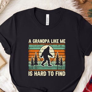 Peut inclure: T-shirt noir avec un graphique rétro d'un Bigfoot debout dans une forêt avec le texte "A Grandpa Like Me Is Hard To Find".