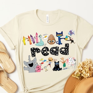 May include: A beige t-shirt with a colorful graphic of characters from the book "The Book With No Pictures". The graphic includes the words "I love to read" and features a variety of characters, including a blue cat, a brown egg, and a pink pig.