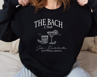Può includere: Felpa nera con testo bianco che recita "The Bach Club Est. 2023" e una grafica di tre cocktail. Il testo "John's Bachelorette Scottsdale, Arizona" è anche stampato sulla felpa.