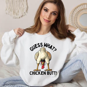 a woman sitting on a bed wearing a white shirt with a chicken saying guess what