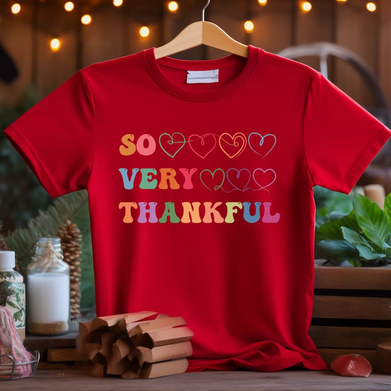 May include: Red t-shirt with the text "SO VERY THANKFUL" in rainbow colors with hearts above the text.