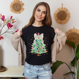 Könnte beinhalten: Ein schwarzes T-Shirt mit einem farbenfrohen Weihnachtsbaum-Design. Der Baum ist mit rosa, grünen, goldenen und weißen Ornamenten geschmückt. Eine Schleife im Leopardenmuster sitzt oben auf dem Baum.