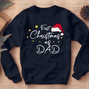 Può includere: Una felpa blu navy con il testo "First Christmas as DAD" in bianco con un grafico di cappello di Babbo Natale rosso e stelle gialle.