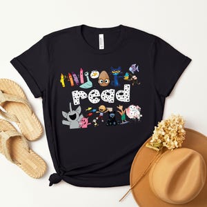May include: Black t-shirt with a graphic of colorful crayons, a cat, an egg, and other characters. The text "Read" is in white with black polka dots.