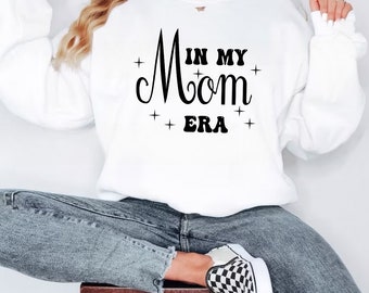 May include: White sweatshirt with black text that reads "In My Mom Era" with stars on either side of the text.