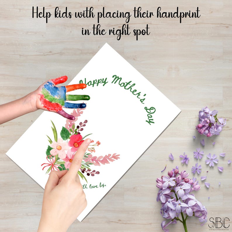 K&ouml;nnte beinhalten: Eine wei&szlig;e Karte mit dem Text "Happy Mother's Day" in gr&uuml;ner Schrift. Eine Aquarell-Blumenzeichnung befindet sich in der linken unteren Ecke. Ein Handabdruck eines Kindes befindet sich in der linken oberen Ecke. Die Karte liegt auf einer Holzoberfl&auml;che.