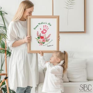 K&ouml;nnte beinhalten: Ein gerahmter Druck mit einem rosa Handabdruck und einem floralen Design. Der Text lautet "Happy Mother's Day Handmade with love from Sophia xo".