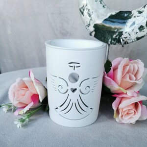 May include: A white ceramic candle holder with an angel design cut out. The angel has wings, a halo, and a heart. Pink roses are arranged around the candle holder. The candle holder is cylindrical.