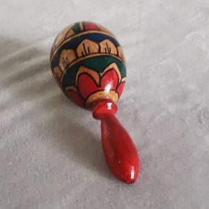 May include: A wooden maraca with a red handle and a colourful painted design. The design features red, yellow, green, and blue colours.