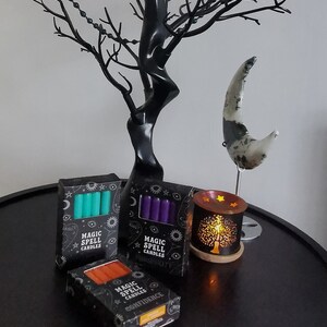 May include: Three boxes of magic spell candles with different colours and scents. The boxes are black with gold lettering and feature a crescent moon, stars, and a sun. The candles are teal, purple, and orange. The boxes are on a dark brown table with a black tree and a white crescent moon sculpture.