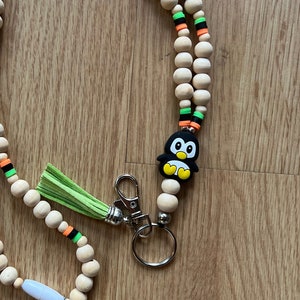 Op de afbeelding: Een houten kralenkoord met een groene kwast, een pinguïnbedel en een metalen sleutelhanger. De kralen wisselen elkaar af in de kleuren oranje, zwart en groen. Het koord heeft de tekst "Ms. McEachern's Math Expert" in witte letters.