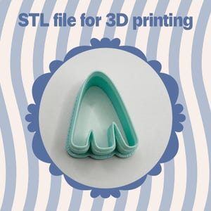 Puede incluir: Un cortador de galletas de plástico azul claro con forma de letra A estilizada, sobre un fondo blanco dentro de un marco festoneado azul. El texto "STL file for 3D printing" está en la parte superior.