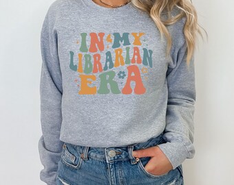 Peut inclure: Un sweat-shirt gris avec le texte "In My Librarian Era" en lettres rétro colorées.