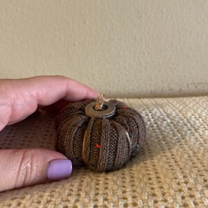 May include: A brown knitted pumpkin with a small metal ring and twine accents. The pumpkin is sitting on a beige knitted surface.