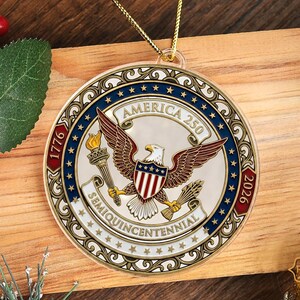 May include: A round, gold-colored ornament with a patriotic design. The ornament features an eagle with outstretched wings, the text "AMERICA 250", and the years "1776" and "2026". The words "SEMIQUINCENTENNIAL" are also present.