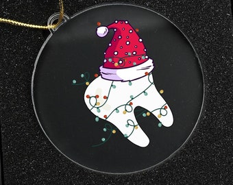 May include: A clear, round Christmas ornament featuring a cartoon tooth wearing a Santa hat and string lights. The ornament is displayed in a black gift box. The box next to it has "Merry Christmas" in red script.
