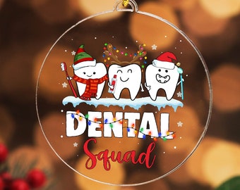 May include: Clear, round Christmas ornament with a Dental Squad theme. Features three cartoon teeth wearing festive hats and holding dental tools. The words "Dental Squad" are written in white and red with Christmas lights.