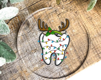 May include: Clear, round Christmas ornament featuring a tooth decorated with reindeer antlers, holly, and colorful Christmas lights. The ornament hangs from a gold string and is set against a rustic wooden background. A festive dental-themed holiday decoration.