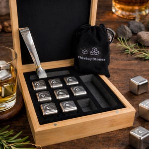 May include: A wooden box set containing whiskey stones, tongs, and a small black bag. The box is open, revealing the stones arranged in a black felt-lined interior. Several stainless steel cubes are outside the box.